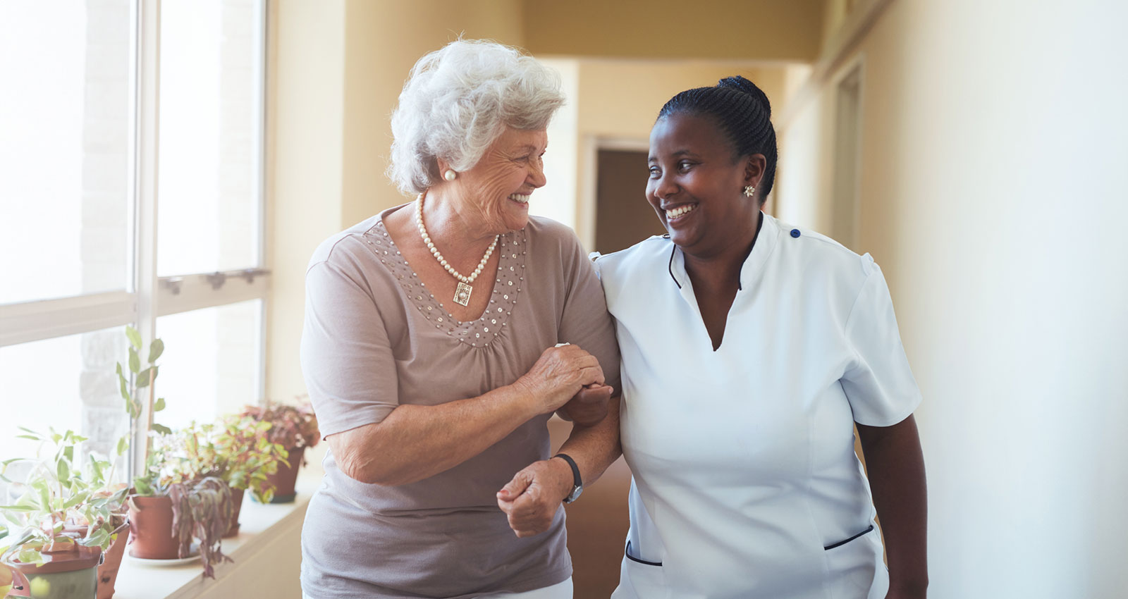 Portrait of smiling home caregiver and senior woman walking together through a corridor. Healthcare worker taking care of elderly woman.